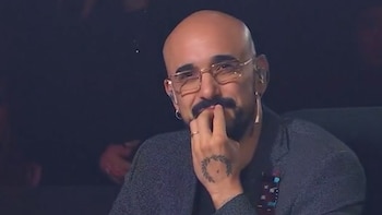 Un hombre calvo con barba y gafas doradas sonríe levemente mientras se toca los labios con la mano derecha, donde se ve un anillo y un tatuaje de laurel