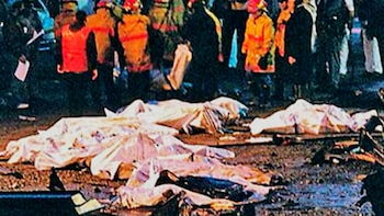 Fotografía granulada y oscura que muestra varios cuerpos cubiertos con sábanas blancas en el suelo. Al fondo, figuras de personas, algunas con chalecos naranjas y cascos