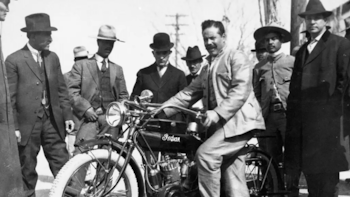 Fotografía en blanco y negro de Francisco Villa sentado en una motocicleta Indian, rodeado por un grupo de hombres vestidos con trajes y sombreros
