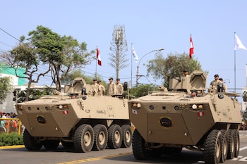 Dos vehículos blindados de ocho ruedas color arena con militares a bordo en un desfile. Banderas de Perú y Corea del Sur. Público en la calle