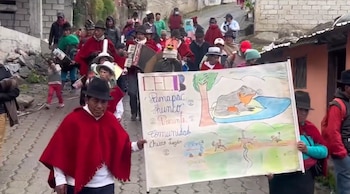 Un grupo de personas vestidas con ponchos rojos y sombreros camina por una calle empedrada sosteniendo un cartel ilustrado y un acordeón