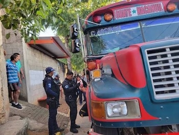 Autobús de colores rojo y azul, agentes uniformados, varias personas, edificios, árboles en una calle