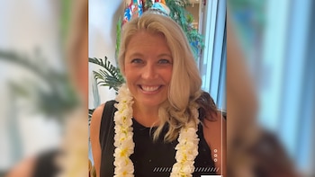 Mujer rubia de ojos azules sonríe a la cámara, lleva un top negro y un collar de flores blancas y amarillas. El fondo borroso muestra plantas verdes