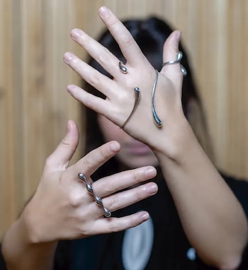 Manos en primer plano que lucen anillos y una pulsera rígida de metal plateado frente al rostro desenfocado de una persona