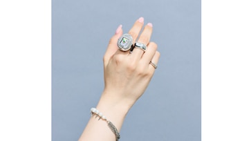Una mano con uñas de color rosa lleva un anillo plateado con la pantalla de un Tamagotchi sobre un fondo gris