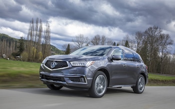 Un SUV 2020 Acura MDX Sport Hybrid gris oscuro se desplaza por una carretera pavimentada, con árboles y colinas verdes bajo un cielo nublado al fondo