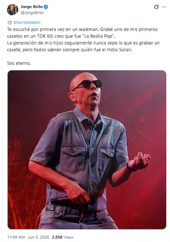 Un hombre calvo con gafas de sol y una camisa de jean está en un escenario oscuro con luces rojas, sosteniendo un micrófono mientras canta