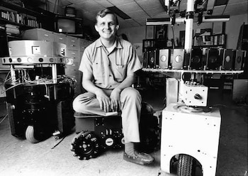 Hombre sonriente sentado sobre un robot con ruedas. Otros dos robots complejos con cámaras y cables en laboratorio. Estanterías, televisores y archivadores en el fondo. Monocromático