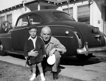 Un hombre y un niño posan de pie junto a un coche clásico estacionado sobre el césped delante de una casa de un piso