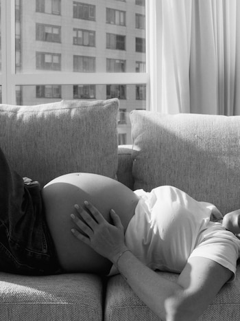 Mujer embarazada acostada en un sofá gris, con la mano sobre su vientre, viste jeans y camiseta blanca, con un edificio visible por la ventana en imagen en blanco y negro