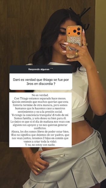 Daniela Celis, de pelo oscuro y blusa blanca, se toma una selfie sonriendo con un celular dorado brillante que muestra un texto blanco en pantalla