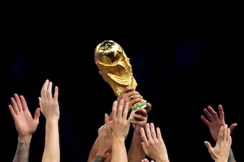 Primer plano del trofeo de la Copa Mundial de la FIFA, de color dorado brillante, siendo sostenido en alto por varias manos sobre un fondo negro