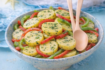 Un cuenco cerámico azul y blanco lleno de rodajas de patata, tiras de pimiento rojo y verde, y perejil picado, con dos cucharas de madera.