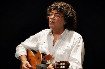 Hombre de cabello rizado y gafas, vestido con camisa blanca, toca una guitarra acústica frente a un fondo negro.