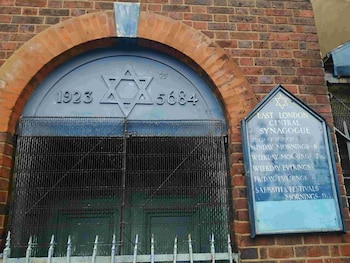Entrada de una sinagoga de ladrillo con un arco. Una puerta metálica con una Estrella de David y fechas. Una reja y un cartel azul a la derecha