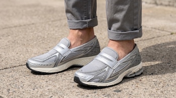 Primer plano de los pies de una persona luciendo zapatillas deportivas tipo mocasín plateadas y grises, con pantalones grises remangados, sobre pavimento.