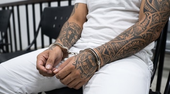 Persona con camiseta y pantalón blancos, esposas de metal en ambas muñecas, y brazos y manos con tatuajes negros de diseños geométricos.