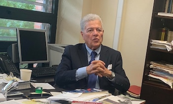 Retrato de Jesús Eduardo Troconis Heredia, un hombre mayor con cabello gris, traje oscuro y corbata azul, sentado en un escritorio con papeles y un monitor