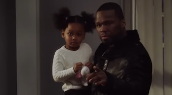 50 Cent con gorra de béisbol negra con el logo de los Yankees, camisa estampada azul y blanca, y pendientes, posa de frente sobre un fondo gris oscuro
