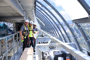 Tres hombres con cascos y arneses de seguridad inspeccionan una pasarela metálica con una estructura de techo arqueado de metal y cristal transparente