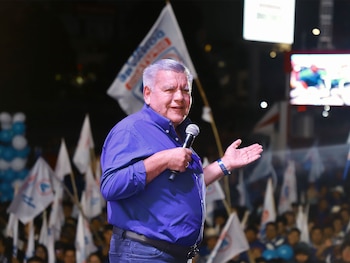 Hombre de mediana edad con camisa azul y micrófono en mano, hablando frente a una multitud y banderas blancas con logo azul en un evento nocturno