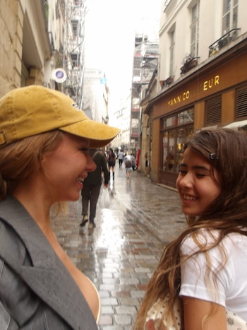 Una mujer con gorra amarilla y una niña sonríen en una calle mojada con locales de fondo