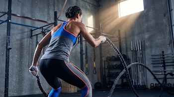 Mujer de espaldas sosteniendo cuerdas pesadas en un gimnasio con paredes de hormigón y equipamiento deportivo.