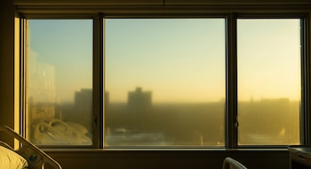 Ventana triple en una habitación de hospital con filtros que suavizan la luz exterior, mostrando siluetas de edificios y un cielo azul y dorado. Parte de una cama médica es visible.