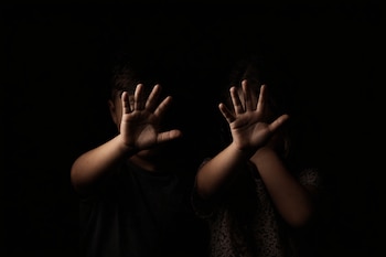 Un niño y una niña aparecen con sus rostros cubiertos por sus manos levantadas, palmas hacia adelante, contra un fondo oscuro y liso.