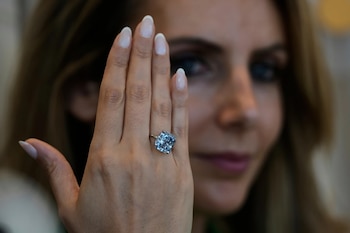 Mano de mujer mostrando un anillo de compromiso con un gran diamante azul rectangular. Uñas con manicura francesa. Rostro borroso de la mujer en segundo plano