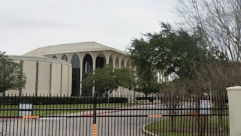 Vista exterior de un edificio moderno color crema con arcos y una fachada curva, detrás de una valla de seguridad negra. Hay árboles y césped verde
