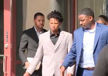 Karmelo Anthony, un joven con traje gris claro y cabello rizado, es escoltado por dos hombres en traje oscuro al entrar o salir de un edificio