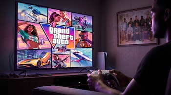 Persona jugando un videojuego en una televisión con el título Grand Theft Auto, un control, una consola, y una foto militar enmarcada en la pared