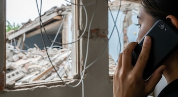 Persona de lado con teléfono móvil negro en la oreja, cabello oscuro. Fondo de casa destruida con paredes caídas, escombros, cables sueltos.