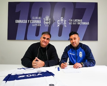Dos hombres en una mesa. Uno firma un documento. Una camiseta de fútbol blanca y azul está sobre la mesa. Un logo del club y texto se ven en la pared