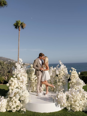Pareja besándose sobre plataforma blanca con arreglos florales blancos. Detrás se ven el mar, la costa montañosa, una palmera y una luna