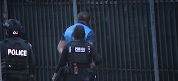 Dos agentes con chalecos que dicen 'POLICE' y 'FEDERAL AGENT' escoltan a un hombre vestido de azul frente a una reja de metal