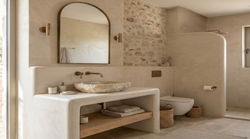 Interior de un baño moderno con lavabo de piedra natural, encimera de estuco, grifos de bronce, espejo arqueado, pared de piedra y ducha abierta.