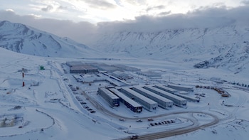 Vista desde la altura de varios edificios modulares alineados, estacionamientos con vehículos y caminos rodeados por montañas con nieve