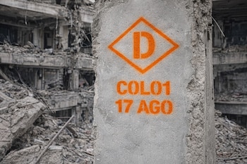Columna de concreto gris con la letra D, COL01 y 17 AGO en esténcil naranja, frente a un edificio demolido con escombros.