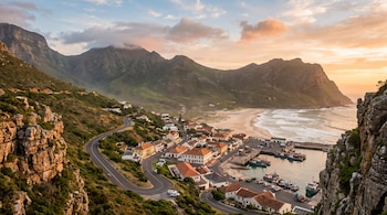 Pueblo costero con casas, puerto con barcos, playa arenosa, carretera, montañas cubiertas de vegetación y acantilados rocosos bajo un cielo con nubes.
