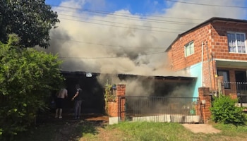 Una densa humareda gris sale de una casa de ladrillos vistos mientras dos personas observan desde el frente