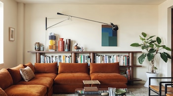 Salón con sofá seccional naranja, estantería de madera llena de libros y objetos, un cuadro abstracto azul y verde, y una planta grande en maceta blanca.