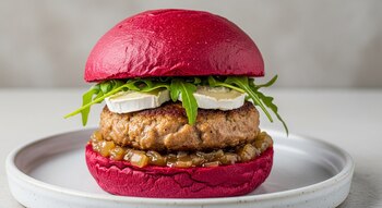 Hamburguesa gourmet de cordero con pan de remolacha fucsia, medallón dorado, queso de cabra, rúcula y chutney de cebolla, servida en un plato blanco.