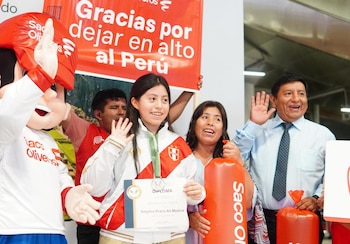 Una joven con casaca deportiva, medalla al cuello y un diploma, rodeada por dos adultos, una botarga y carteles rojos de felicitación