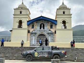 Una iglesia colonial de dos torres con fachada rosada y azul, flanqueada por una reja blanca. Un vehículo policial gris está estacionado frente a sus escaleras, con personas cerca