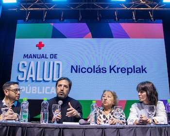 Cuatro personas en una mesa con micrófonos y botellas. Detrás, una pantalla exhibe el título "Manual de Salud Pública" con una cruz roja y el nombre "Nicolás Kreplak"