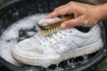 Una mano sostiene un cepillo de madera con cerdas claras, frotando la parte superior de un tenis blanco sucio en un recipiente negro con abundante espuma de jabón.