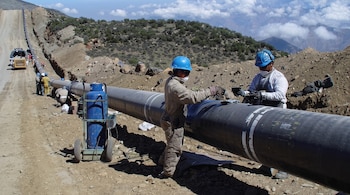 Dos personas con cascos azules y mascarillas trabajan junto a una tubería negra y un cilindro de gas azul en un terreno montañoso.