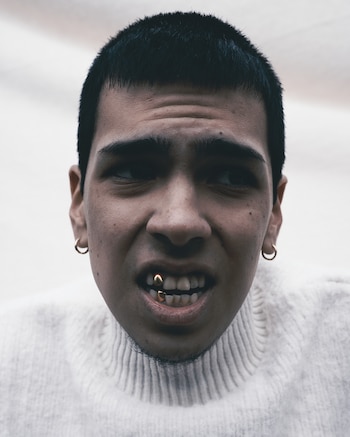 Primer plano de un joven con cabello corto oscuro, suéter blanco de cuello alto, aretes dorados de aro y dos dientes con fundas doradas